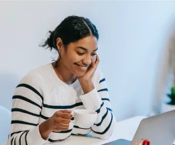 A girl at a laptop, smiling, drinking a cup of coffee